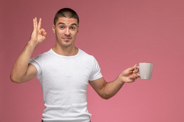 Front view young male in white t-shirt posing and holding cup of coffee on pink background  