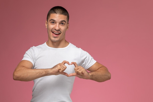 Front view young male in white t-shirt showing heart sign on pink background  