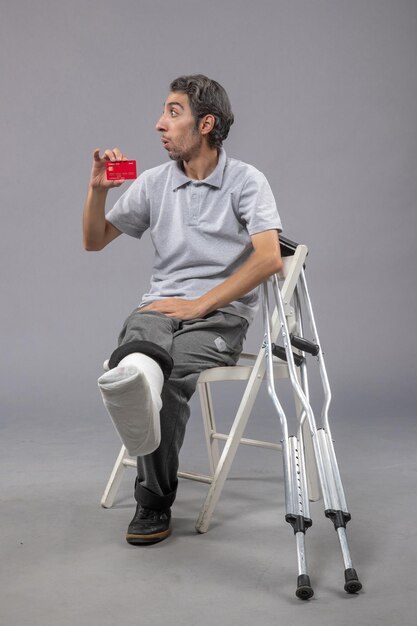 Front view young male with broken foot and bandage holding bank card on grey wall accident twist foot human pain male