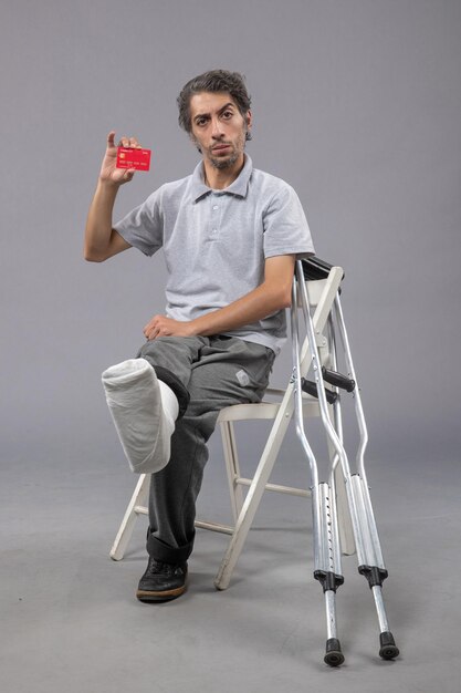 Front view young male with broken foot and bandage holding bank card on grey wall pain twist leg foot human male