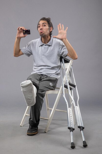 Front view young male with broken foot and bandage holding black bank card on the grey wall twist male accident foot pain