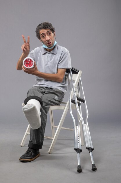 Front view young male with broken foot and bandage holding clock on grey desk twist leg foot human pain male