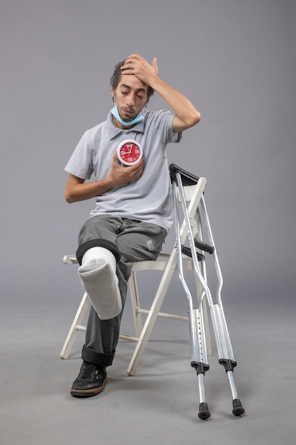 Front view young male with broken foot and bandage holding clock on a grey wall pain twist leg foot human male