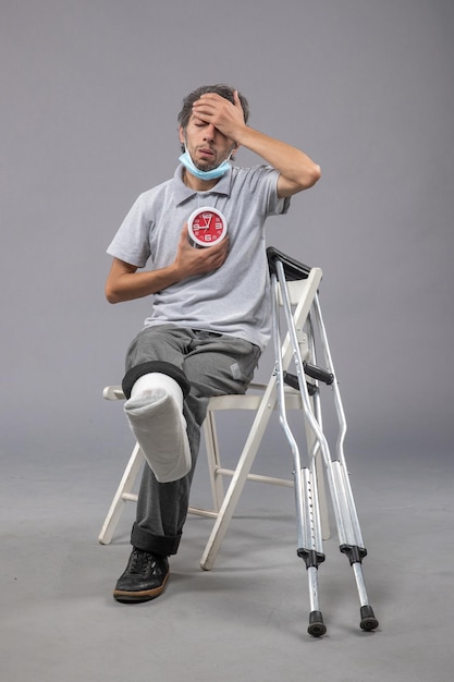 Front view young male with broken foot and bandage holding clock on grey wall pain twist leg foot human male