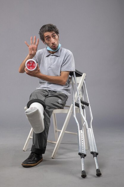 Front view young male with broken foot and bandage holding clock on the grey wall twist leg foot human pain male