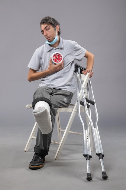 Front view young male with broken foot and bandage holding clock on a grey wall twist leg pain male foot human