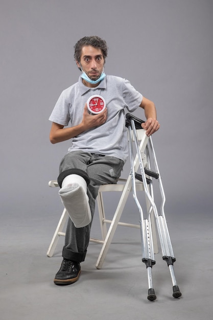 Front view young male with broken foot and bandage holding clock on the grey wall twist leg pain male foot human