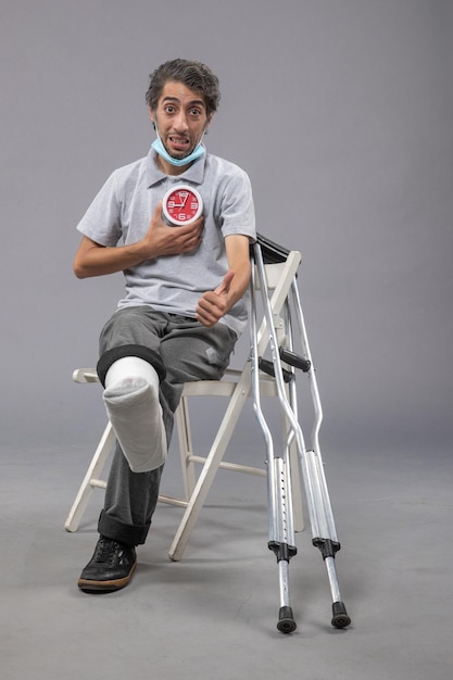 Front view young male with broken foot and bandage holding red clock on grey wall twist leg human pain male foot