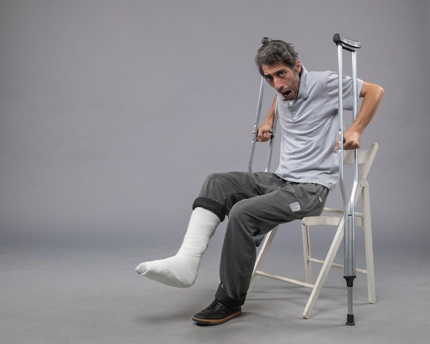 Free photo front view young male with broken foot trying to stand up holding crutches on grey wall foot broken pain twist accident leg