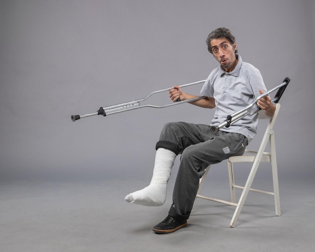 Front view young male with broken foot trying to stand up holding crutches on grey wall foot broken pain twist accident legs