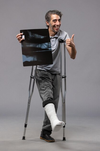 Front view young male with broken foot using crutches and holding his x-ray on the grey wall disable accident foot twist broken pain