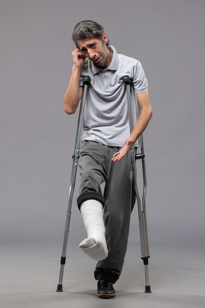 灰色の壁に足が折れたため松葉杖を持った若い男性の正面図足が折れた事故の足を無効にする