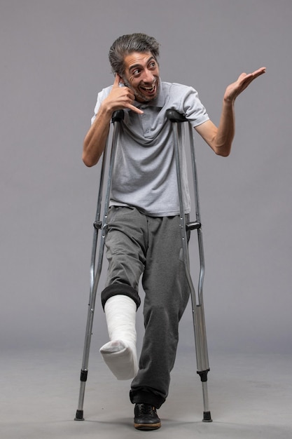 Бесплатное фото Вид спереди молодого мужчины с костылями из-за сломанной ноги на серой стене, отключившей сломанную ногу в результате несчастного случая