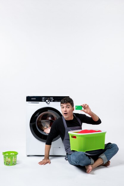 Front view of young male with green basket full of clothes holding bank card on white wall