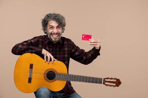 Front view of young male with guitar holding bank card on a pink wall