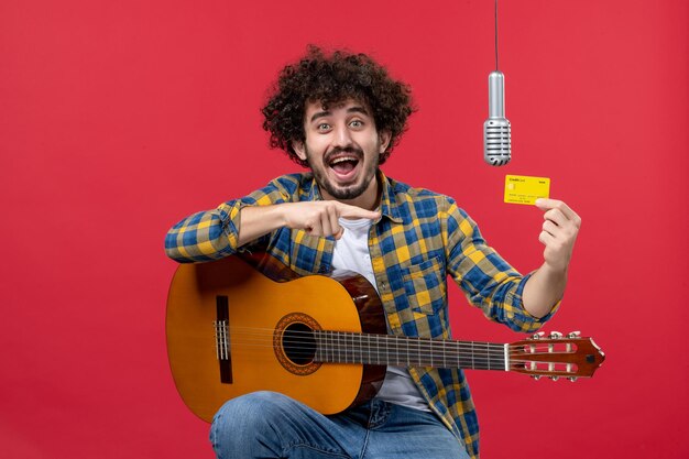 Front view young male with guitar holding bank card on the red wall band singer live performance musician concert money color