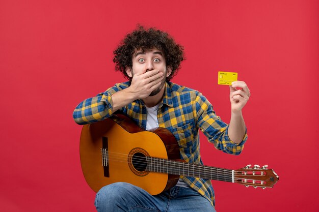 Front view young male with guitar holding bank card on red wall color performance applause musician live concert band money