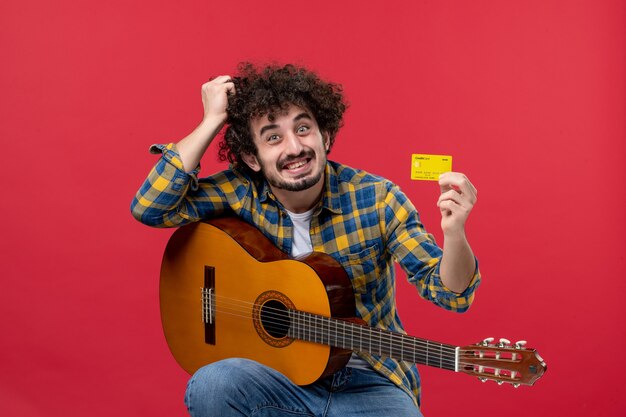 Front view young male with guitar holding bank card on red wall color performance musician live concert band money