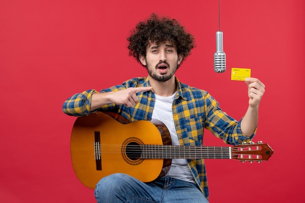 Front view young male with guitar holding bank card on a red wall live color performance musician concert band money