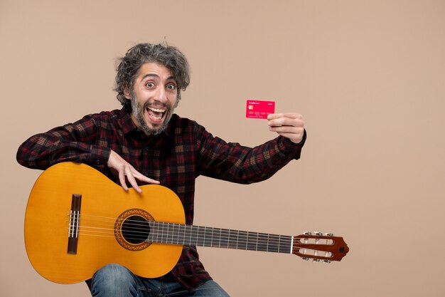 Front view of young male with guitar holding red bank card on pink wall