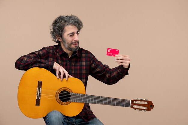 Front view of young male with guitar holding red bank card on pink wall