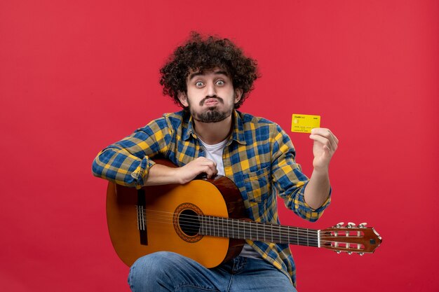 Вид спереди молодой мужчина с гитарой, держащий желтую банковскую карту на красной стене, цветное выступление, аплодисменты, живой концерт музыканта