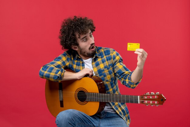 赤い壁に黄色の銀行カードを保持しているギターを持つ正面図若い男性カラーパフォーマンス拍手ミュージシャンライブコンサート