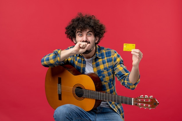 Front view young male with guitar holding yellow bank card on a red wall color performance concert applause musician live music