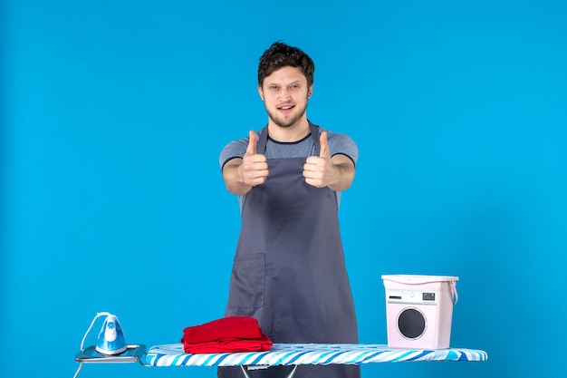 Front view young male with ironing board and clothes on a blue background color housework laundry cleaning washing machine