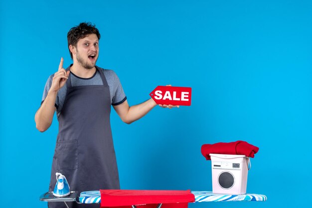 Front view young male with ironing board and sale in his hands on blue background cleaning laundry iron washing machine housework colors