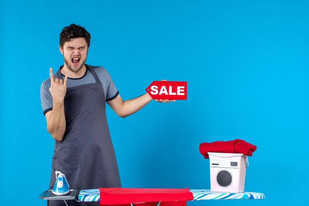 Front view young male with ironing board and sale in his hands on blue floor cleaning laundry washing machine housework color iron