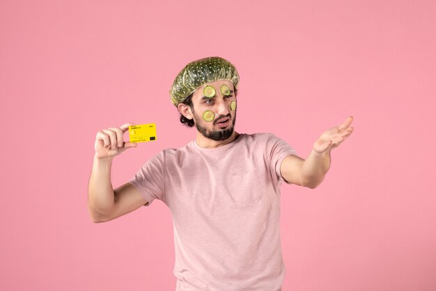 ピンクの背景に銀行カードを保持している彼の顔にマスクを持つ若い男性の正面図