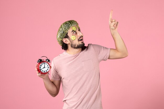 вид спереди молодой мужчина с маской на лице, держащий часы на розовом фоне