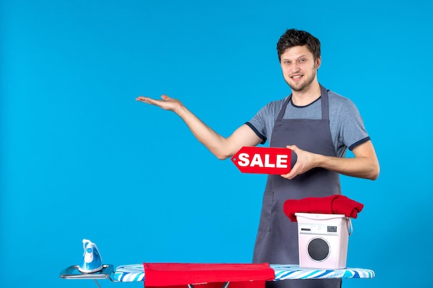 Вид спереди молодой мужчина с красной распродажей, пишущий в его руках на синем фоне, прачечная, утюг, покупка, стиральная машина, чистка
