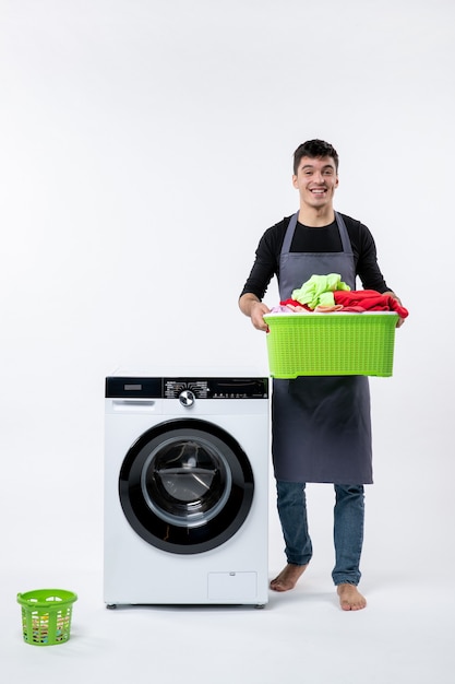 Front view of young male with washer and dirty clothes inside basket on white wall