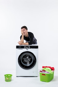 Vista frontale del giovane maschio con lavatrice e vestiti sporchi sul muro bianco
