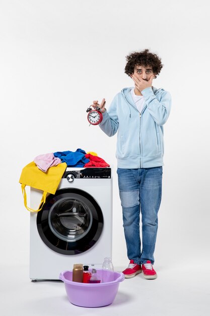白い壁に時計を保持している洗濯機を持つ若い男性の正面図