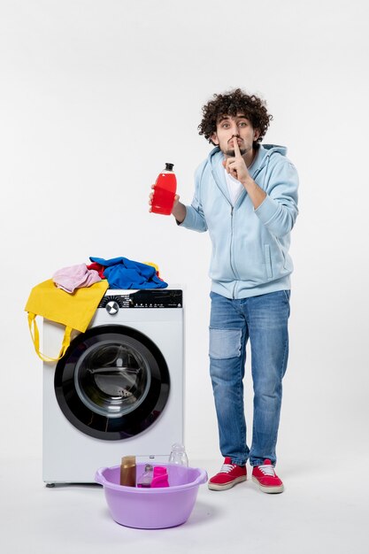 흰 벽에 액체 가루를 들고 있는 세탁기가 있는 젊은 남성의 전면 모습