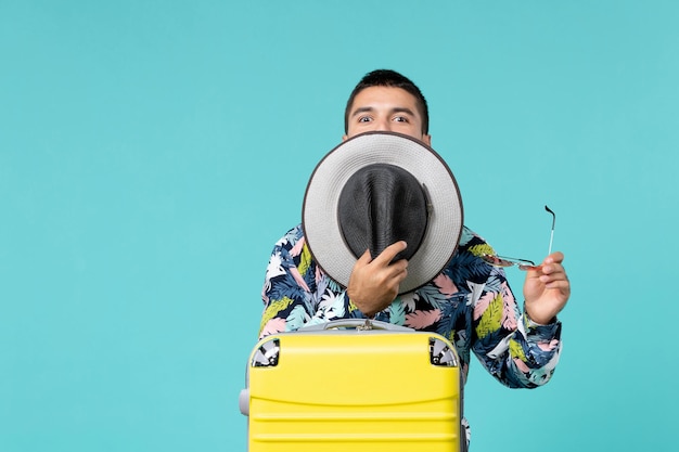 Vista frontale del giovane maschio con la borsa gialla che tiene il cappello e si prepara per il viaggio sulla parete azzurra