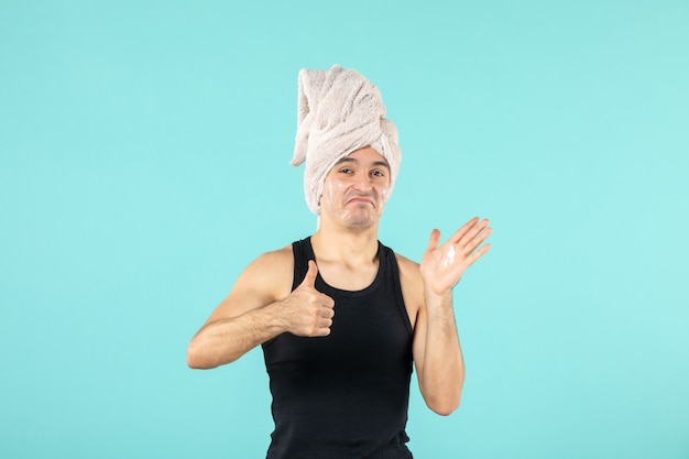 front view of young man after shower applying cream on his face on blue wall