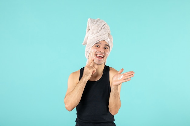 front view of young man after shower applying cream on his face on blue wall