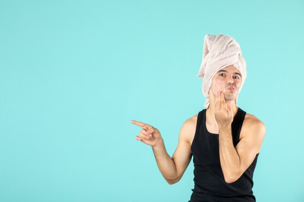 front view of young man after shower applying cream to his face on blue wall