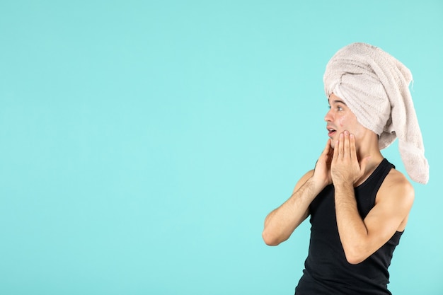 front view of young man after shower applying cream to his face on blue wall
