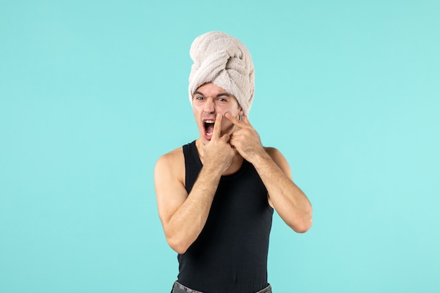 front view of young man after shower trying to squeeze out little pimple on blue wall