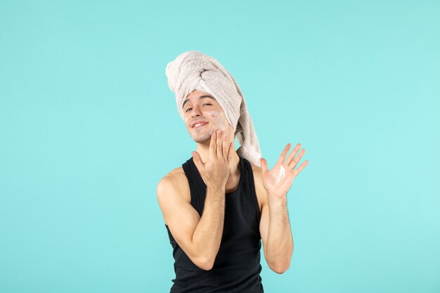 front view of young man after shower with cream on his face on blue wall