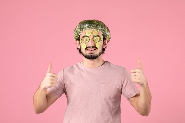 front view of young man applying cucumber mask on his face on pink wall