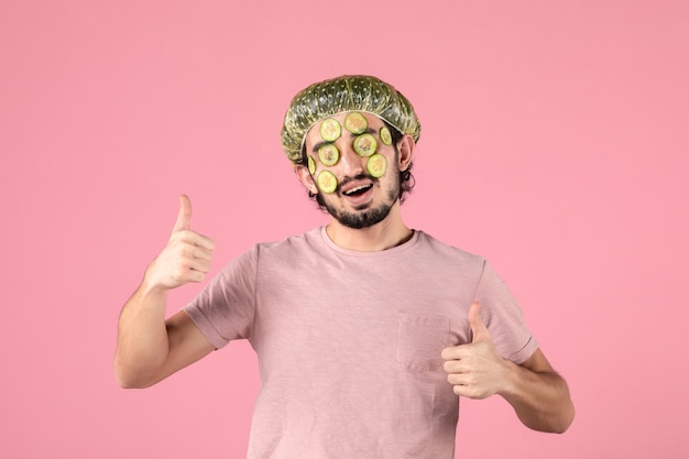 front view of young man applying cucumber mask on his face on pink wall