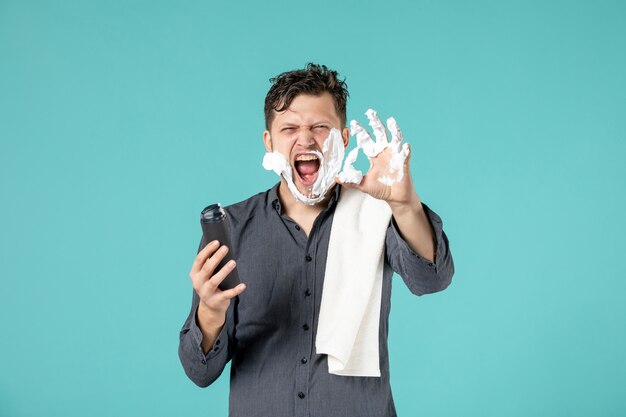 front view of young man applying foam for shaving into his face on blue wall