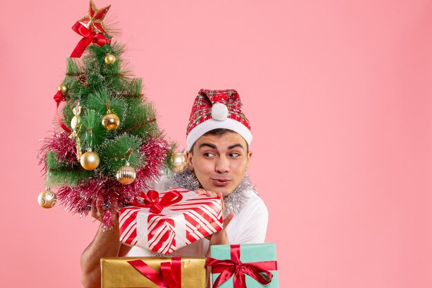 Вид спереди молодого человека вокруг рождественских подарков и праздничной елки на розовой стене