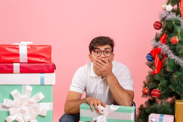 Front view of young man around presents and christmas tree on pink wall